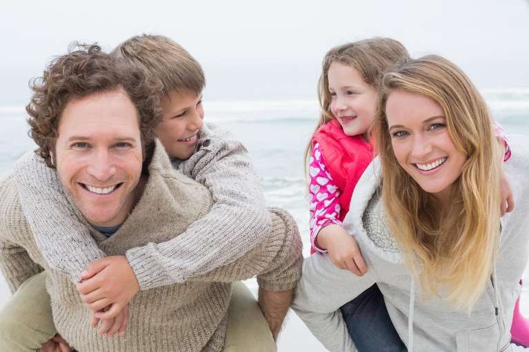Smiling family on the beach with parents giving children piggyback rides during travel.
