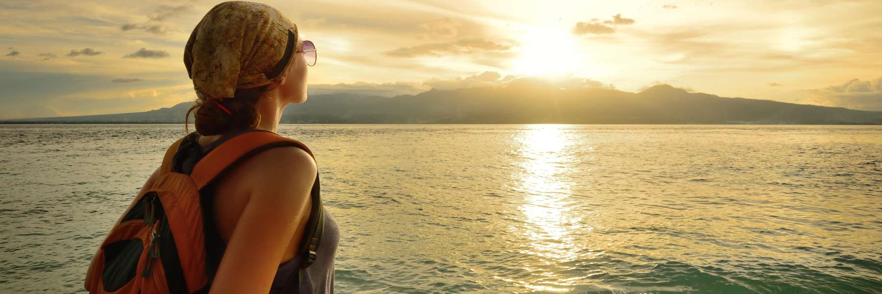Woman with backpack looking at ocean sunset while traveling.